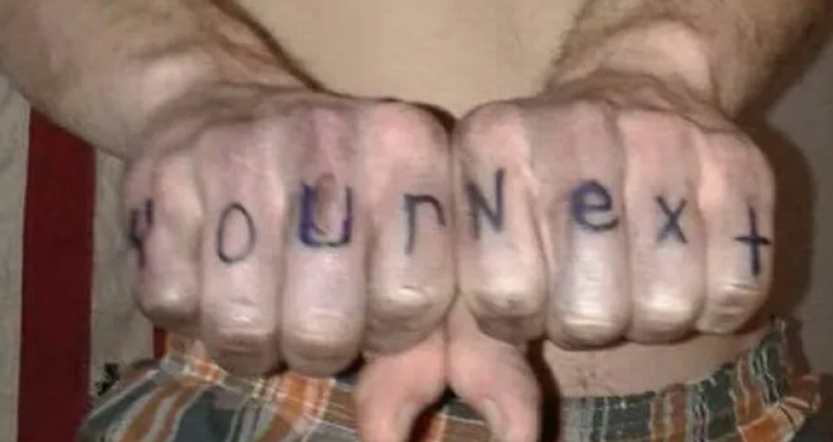 A person with bare arms and plaid shorts has the words "Your Next" written in black marker across the knuckles of both fists, which are held together and facing the camera.
