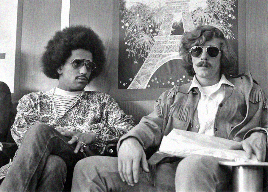 Two men sit side by side wearing sunglasses and stylish clothing, one with an afro and patterned shirt, the other with wavy hair, a mustache, and fringed jacket. Behind them is a poster of the Eiffel Tower with fireworks.