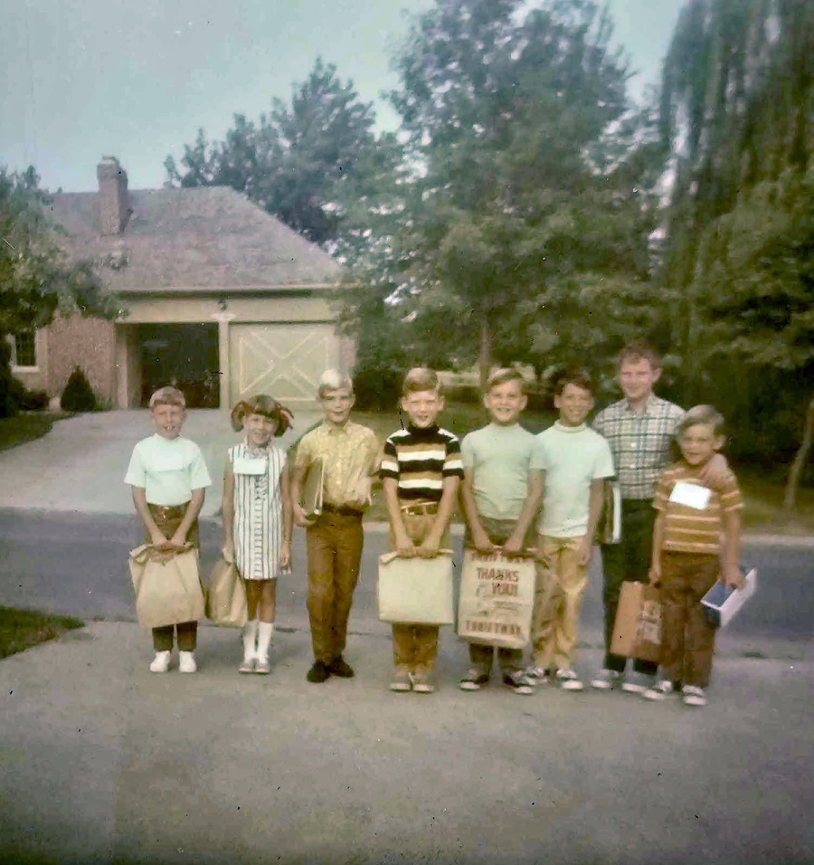 Eight children stand in a row on a suburban street, holding bags and smiling. Trees and a house with a garage are visible in the background. The photo has a vintage, faded look.