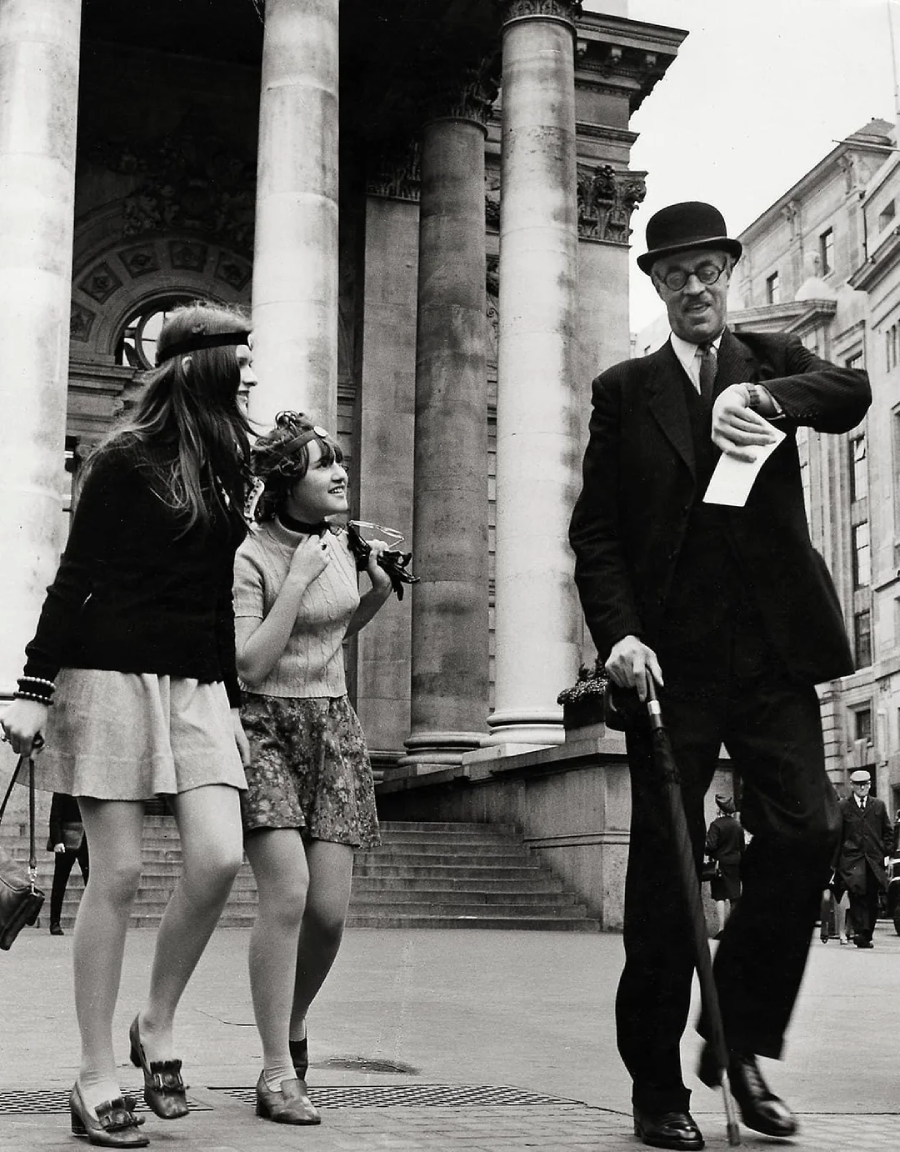 Two young women laugh and follow an older man in a suit, bowler hat, and glasses, who walks with a cane and holds a paper. They stand in front of a grand building with large columns.