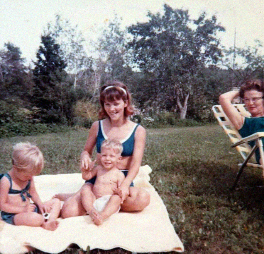 A woman sits on a blanket outside with two young children; another woman sits on a lawn chair nearby. They are in a grassy area with trees, all wearing summer clothes and smiling.