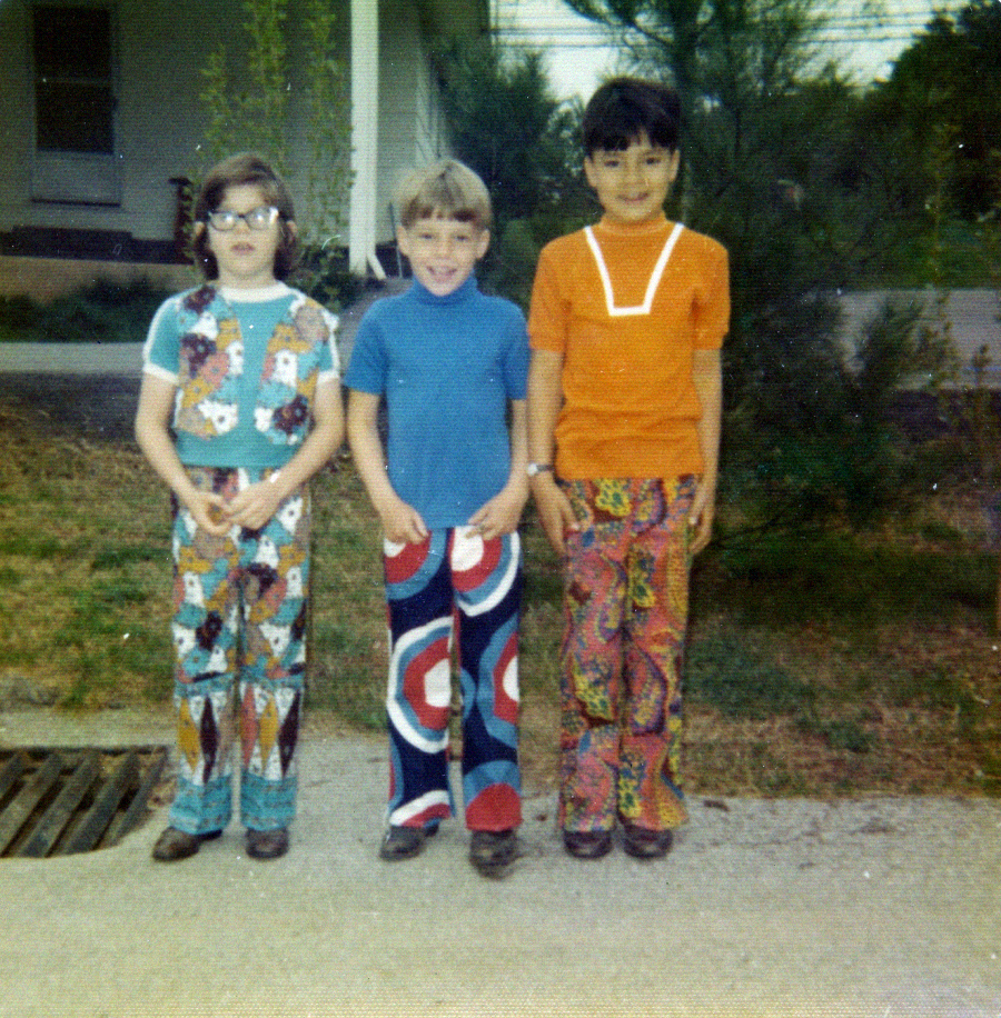 Three children stand side by side outdoors, each wearing colorful 1970s clothing with bold patterns. The background features a house, greenery, and a drain grate on the ground.