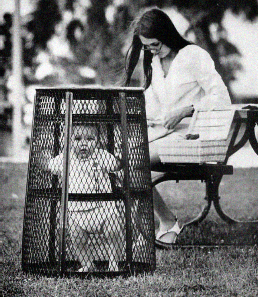 A woman sits on a park bench, smiling and watching a baby who is standing inside a large metal mesh enclosure on the grass. The scene appears to be from a past era, possibly the 1960s or 1970s.