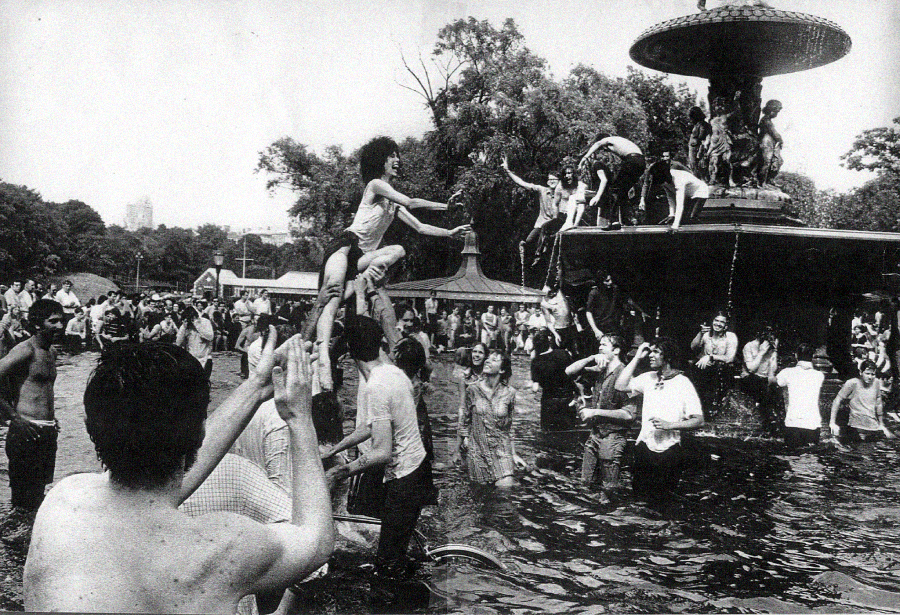 A large group of people play and splash in a city park fountain on a sunny day, with some climbing on the fountain and others helping each other in the water. The scene is lively and joyful.
