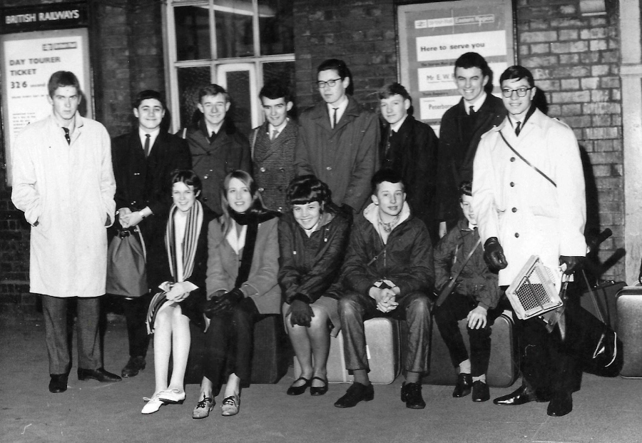 A black-and-white photo of a group of young men and women in coats, sitting and standing on a train station platform with suitcases. Posters and brick walls are visible in the background.