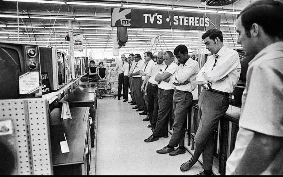 A group of men stand in a line, intently watching television sets on display in a store, under a sign that reads “TV’s - STEREOS.” The scene appears to be from a past decade.