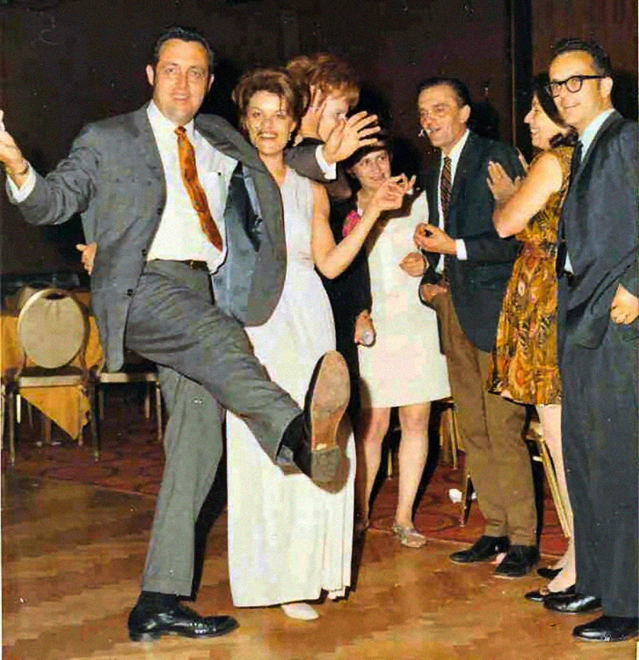 A group of people dressed in formal and semi-formal clothing are dancing and smiling in a line at an indoor event. The man in front lifts his leg enthusiastically, and the others wave and appear joyful.