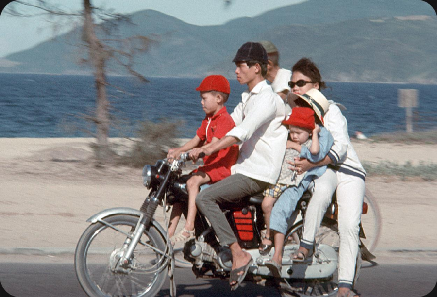 A family of five, including three children, rides together on a motorcycle near a beach. The father drives while the others sit behind him. The background shows water, sand, mountains, and a tree.