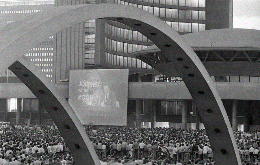 A large crowd gathers at an outdoor plaza, watching a giant screen displaying “Journey to the Moon.” Arching structures frame the scene, with tall modern buildings in the background. The image is in black and white.