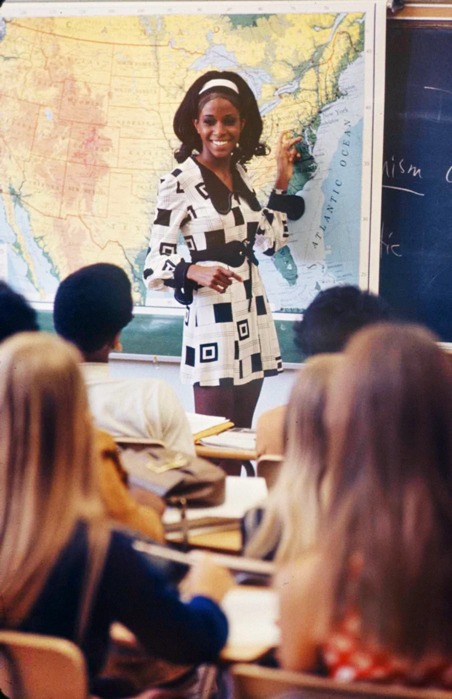 A teacher in a patterned dress stands in front of a U.S. map and blackboard, smiling and pointing at the East Coast, while students sit at desks and listen attentively.