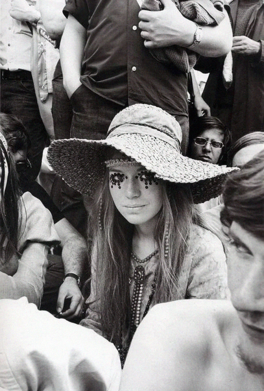 A young woman wearing a large straw hat and beaded face decorations sits among a crowd of people at an outdoor event. The image is black and white and appears to be from the 1960s or 1970s.