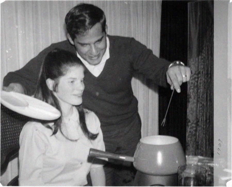 A man and woman prepare fondue together; he holds a fork and she sits smiling. The photo is black and white, with "1969" written in the corner, capturing a casual, joyful moment.