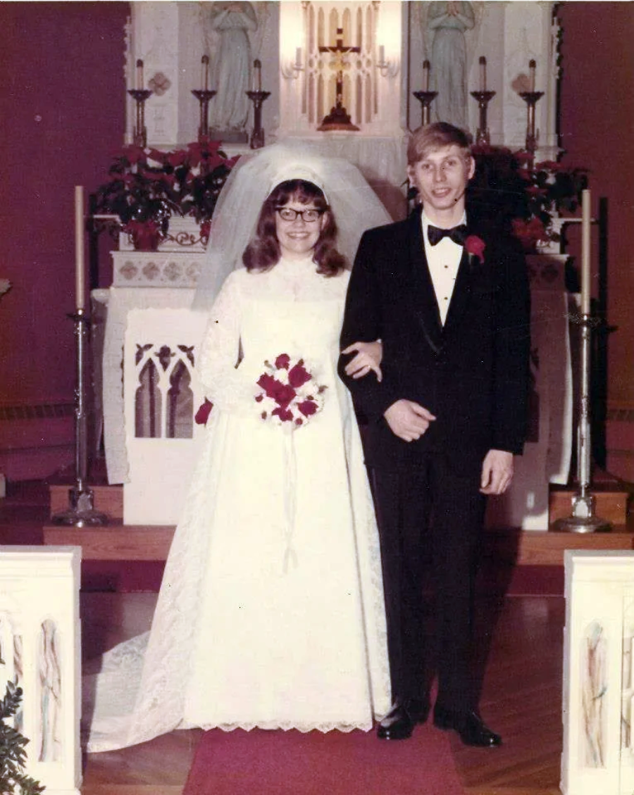 A bride in a white gown and veil stands arm-in-arm with a groom in a black tuxedo in a church decorated with flowers and candles, posing at the altar on their wedding day.