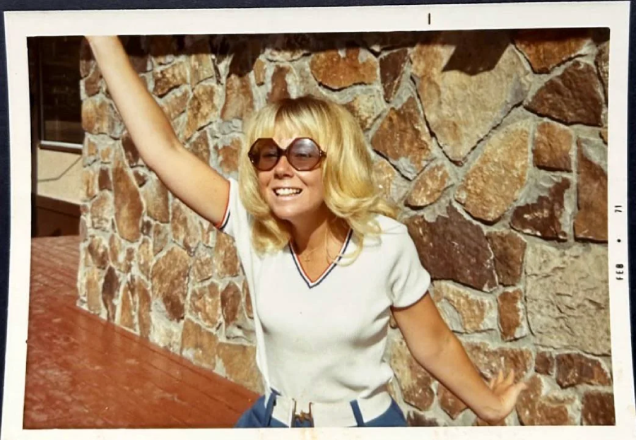 A smiling woman with blonde hair and sunglasses poses with one arm raised in front of a stone wall, wearing a white shirt and blue pants. The photo has a vintage appearance.