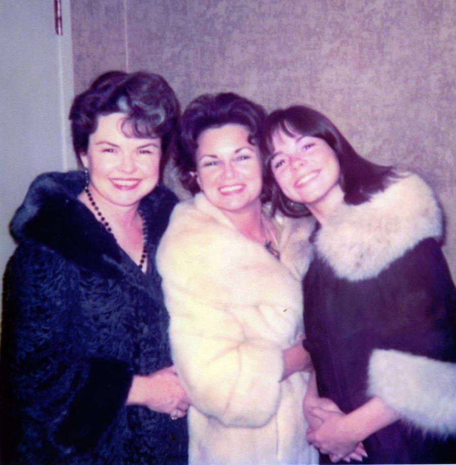 Three women stand closely together, smiling at the camera. They are dressed in elegant fur coats, with a plain wall as the background. The photo appears to be from a past decade, possibly the 1960s or 1970s.
