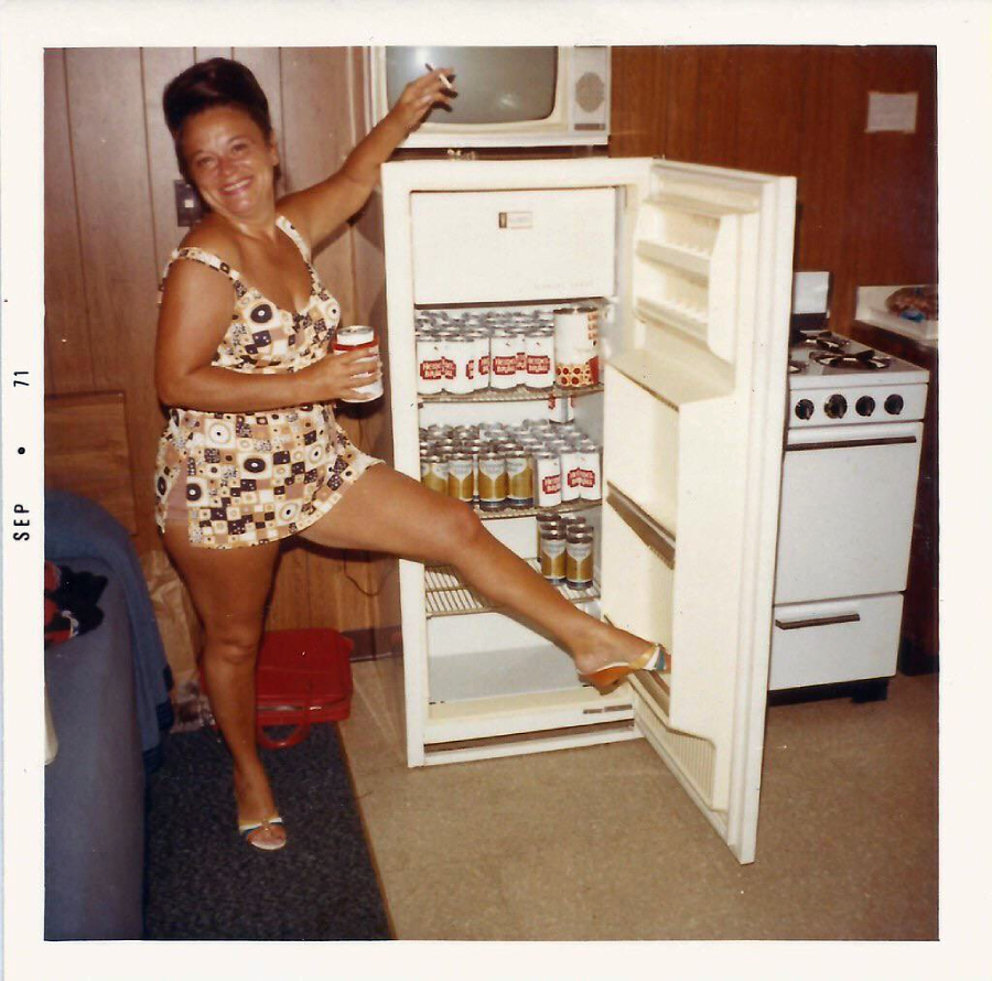 A smiling woman in a patterned swimsuit stands next to an open fridge filled with cans, holding a drink and raising one leg to rest her foot on the fridge door. A small stove and wood-paneled walls are in the background.