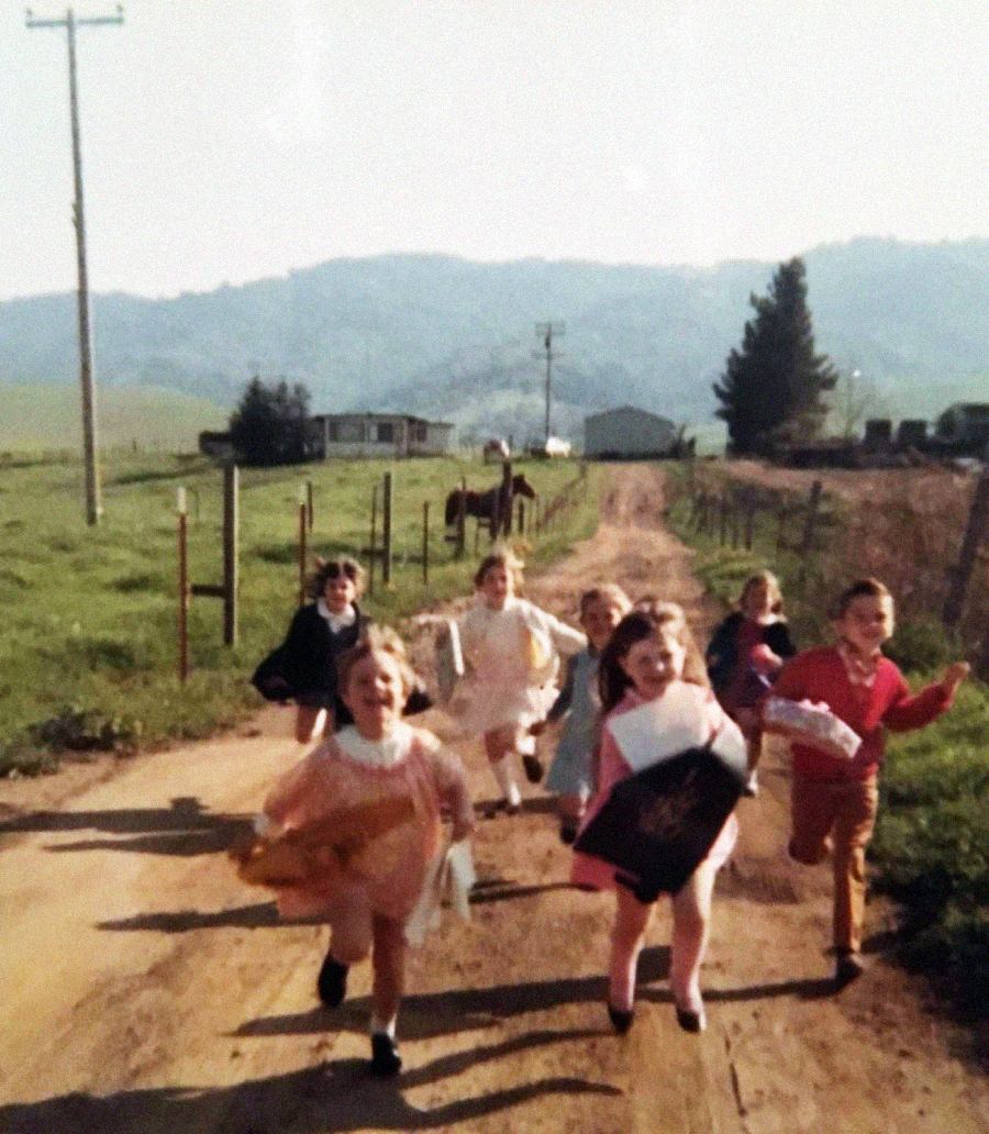 A group of children, dressed in colorful clothes, joyfully run down a dirt path in a rural area, with green fields, a fence, and distant hills in the background on a sunny day.