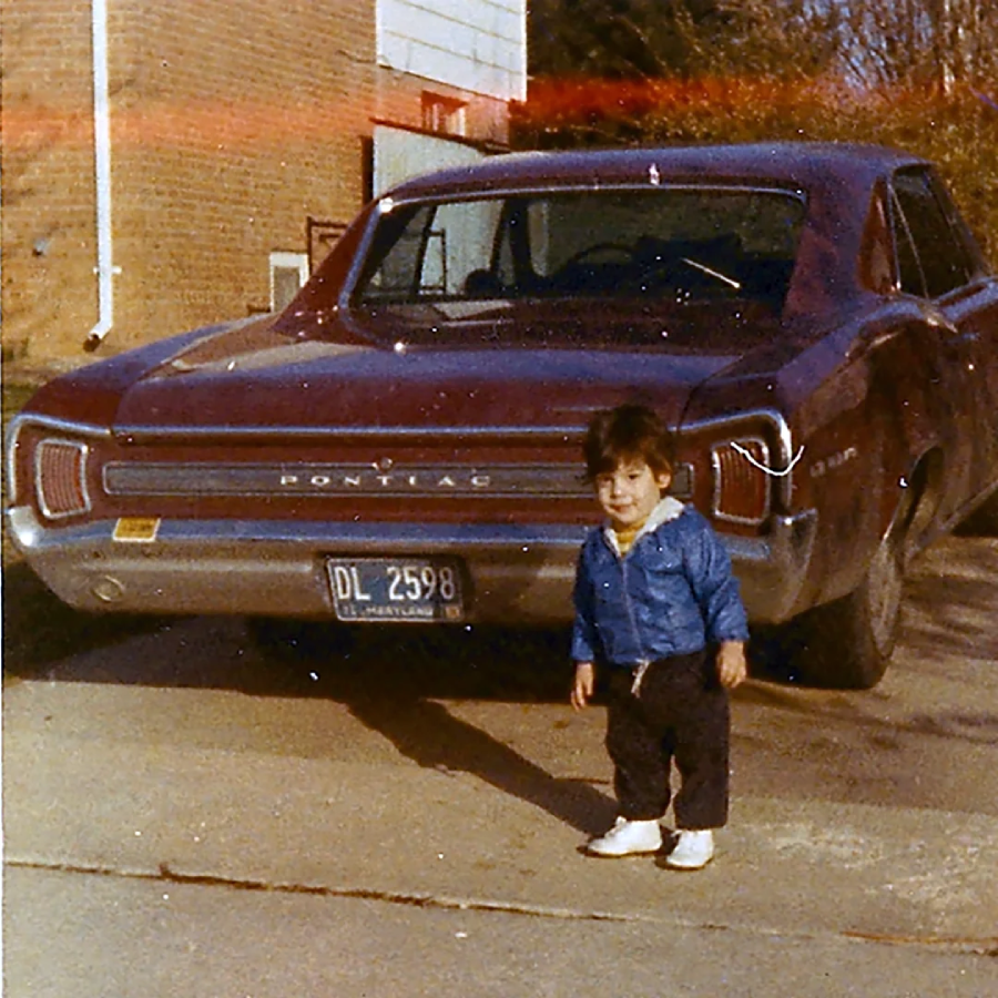 A young child in a blue jacket and white shoes stands on a driveway next to a vintage maroon Pontiac car with a visible license plate, in front of a brick building and some trees.