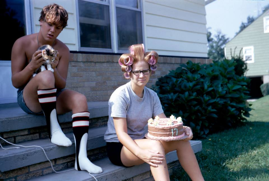 A young man in knee-high striped socks holds a small dog on porch steps beside a woman with pink hair rollers and glasses, who is smiling and holding a birthday cake with the number 17 outside a house.