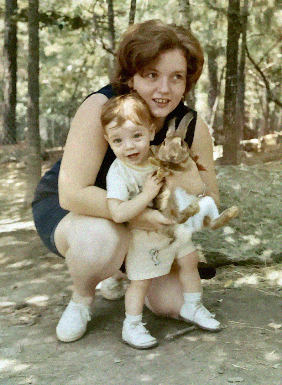 A woman crouches behind a young child holding a brown and white rabbit. They are both smiling outdoors in a wooded area, with trees and a fence in the background.