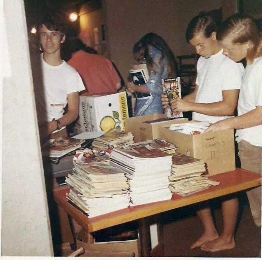 Three young people sort through stacks of comic books on a table, with boxes labeled "comics" nearby. The setting appears to be indoors, possibly at an event or sale. One person stands barefoot, while others look focused.