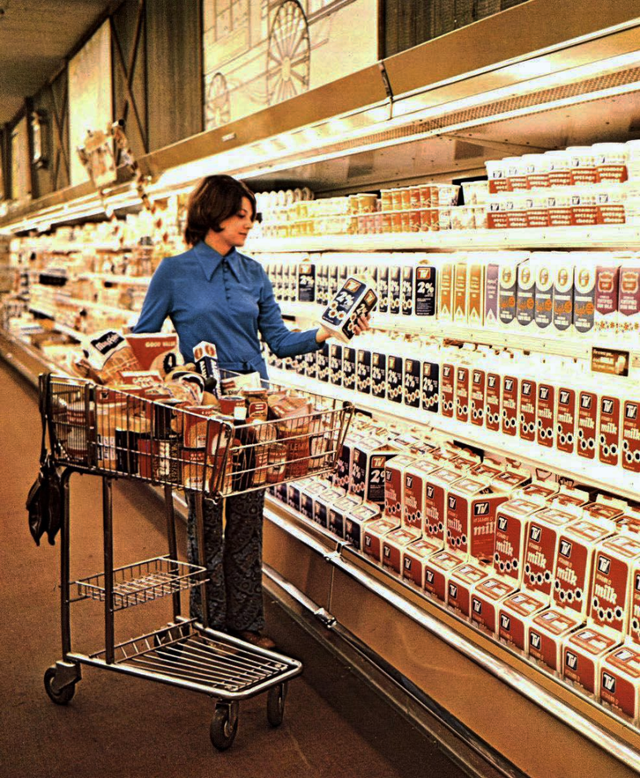 A woman in a blue shirt shops for milk in a supermarket aisle lined with various milk cartons. She holds two cartons and has a full shopping cart beside her. The scene appears to be from the 1970s.
