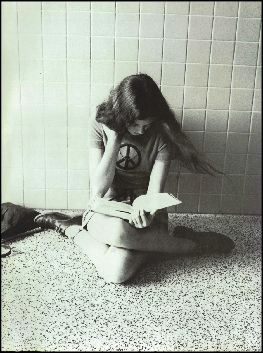 A young woman with long hair sits on the floor against a tiled wall, wearing a shirt with a large peace symbol and reading a book. Her legs are crossed and a bag rests nearby.