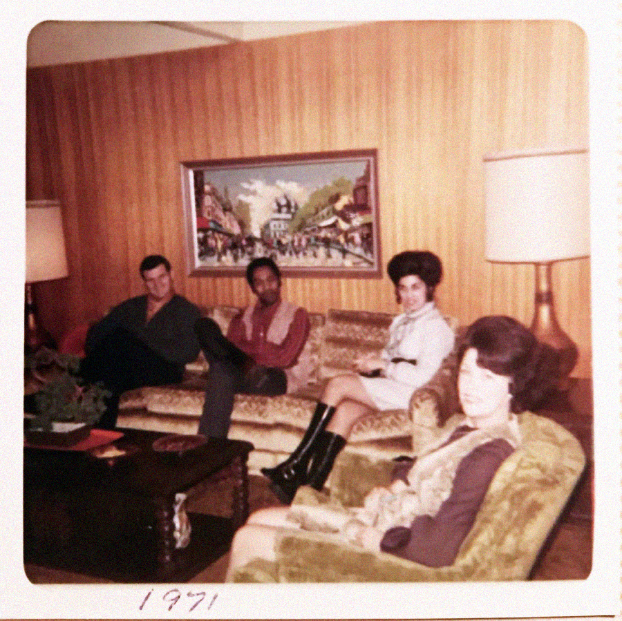 Four people sit in a living room with wood-paneled walls, vintage furniture, and a painting on the wall. Two men and two women are seated on sofas, dressed in 1970s-style clothing. "1971" is written on the photo.