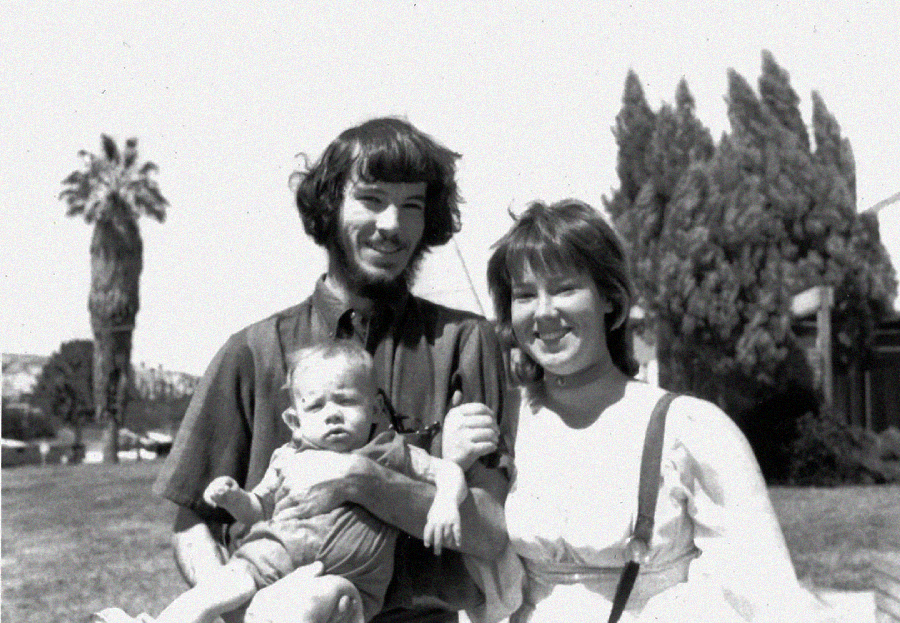 A smiling young couple stands outside holding a baby. The man has medium-length hair and a beard, while the woman has shoulder-length hair. Trees and a palm tree are visible in the background on a sunny day.