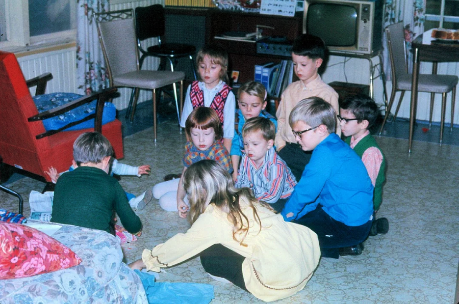 A group of children sits on the floor in a living room, gathered around an unseen object, with retro furniture and a vintage television in the background.