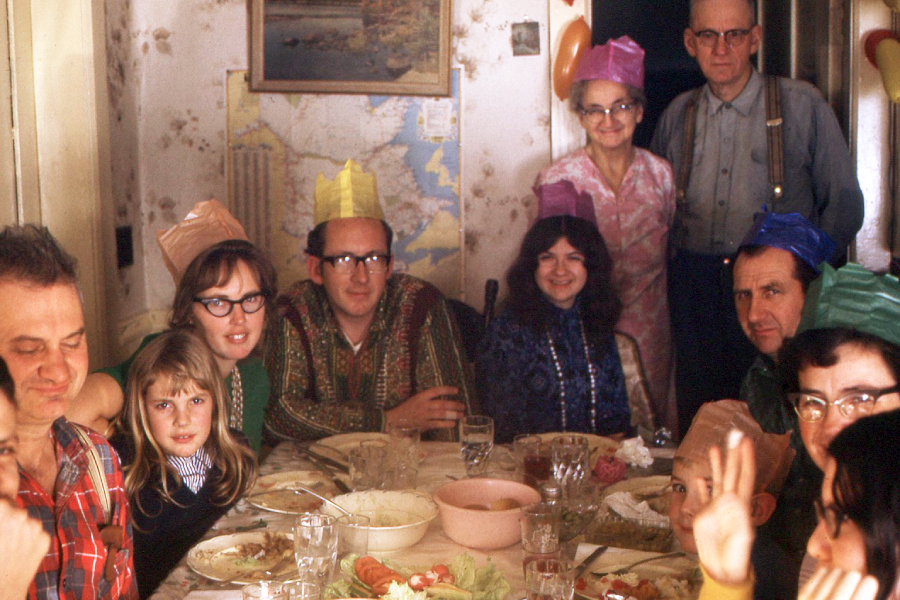 A group of adults and children wearing colorful paper hats sit and stand around a dining table set with food, smiling and posing for a photo in a cozy, decorated room.