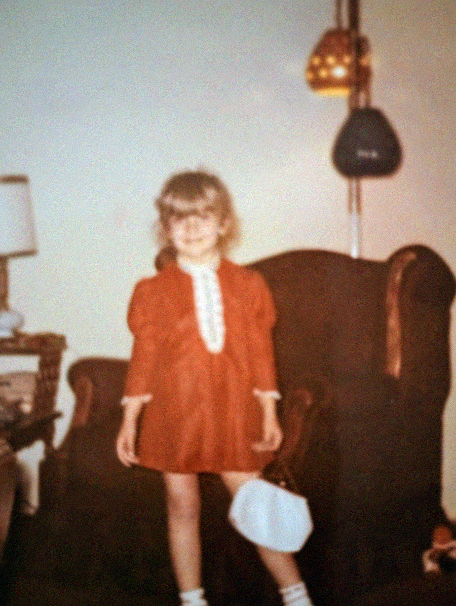 A young girl with blonde hair wearing a red dress and white shoes stands in front of a dark armchair, holding a white handbag. There is a lamp and a side table with a lamp in the background. The photo has a vintage feel.