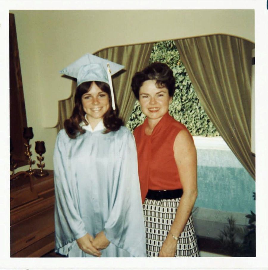 A young woman in a light blue graduation cap and gown stands smiling next to an older woman in a sleeveless red top and patterned skirt. They are indoors, in front of a window with tan curtains.