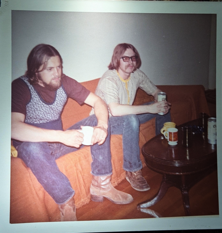 Two men with long hair and beards sit on an orange-covered couch, holding drinks. One wears a patterned vest, the other sunglasses and cowboy boots. A small table with mugs and cans is in front of them.