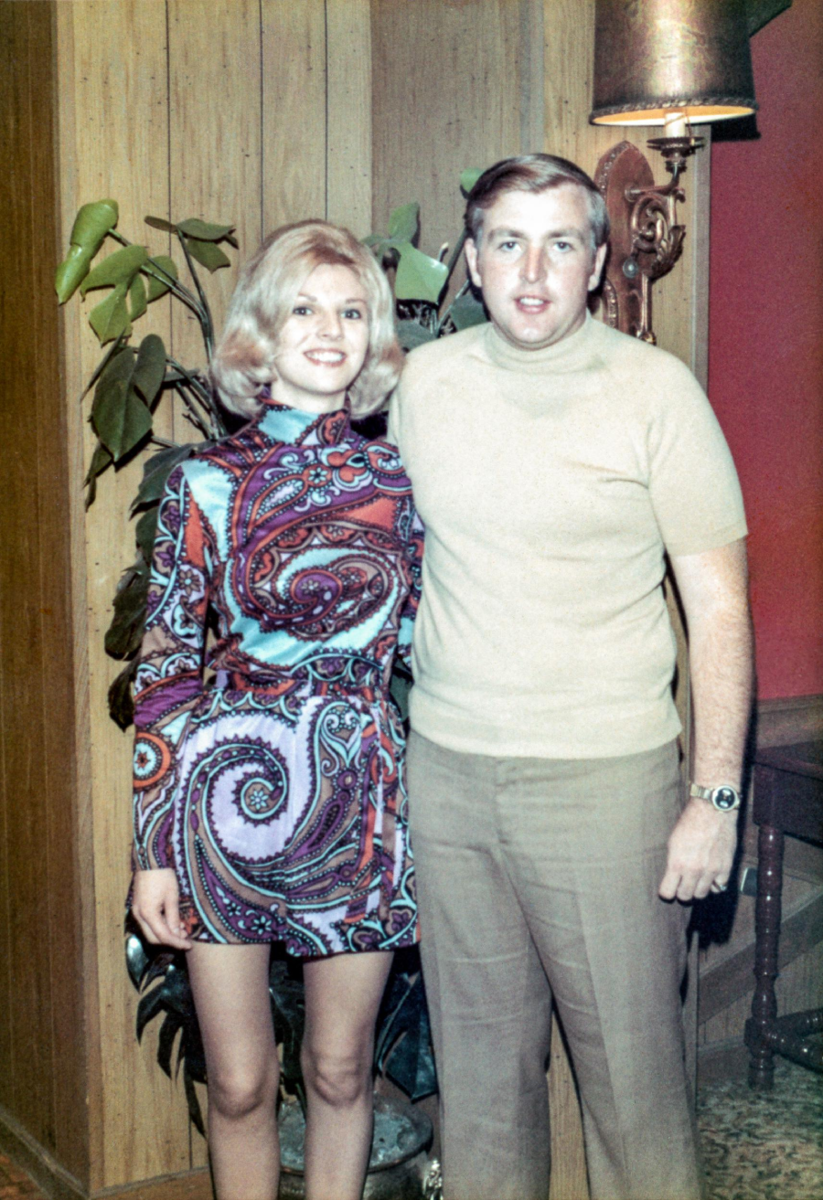 A woman in a colorful patterned dress and a man in a beige turtleneck and brown pants stand together indoors, smiling, in a retro-style room with wood paneling and a potted plant.