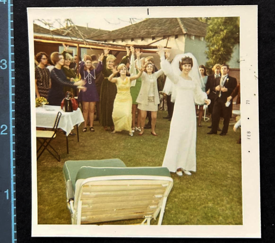 A bride in a white dress and veil smiles while tossing her bouquet to a group of excited women standing behind her at an outdoor gathering, with tables and chairs set up on the lawn.