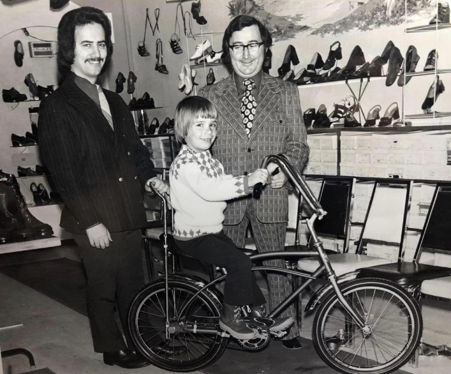 A black-and-white photo of two men in suits standing beside a smiling child sitting on a bicycle inside a shoe store with shelves of shoes in the background.