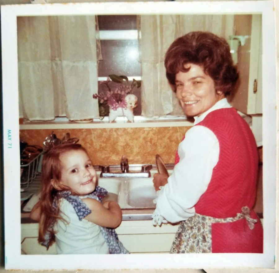 A woman and a young girl smile at the camera while standing at a kitchen sink. The woman wears a red vest and apron; the girl has long brown hair. The kitchen has yellow walls and white curtains. The photo is dated May 1971.