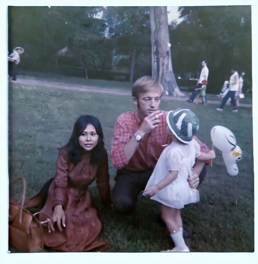 A woman in a long dress sits on grass beside a man in a red shirt, who is kneeling and holding a child in a white dress and sunhat. They appear to be in a park with trees and people in the background.