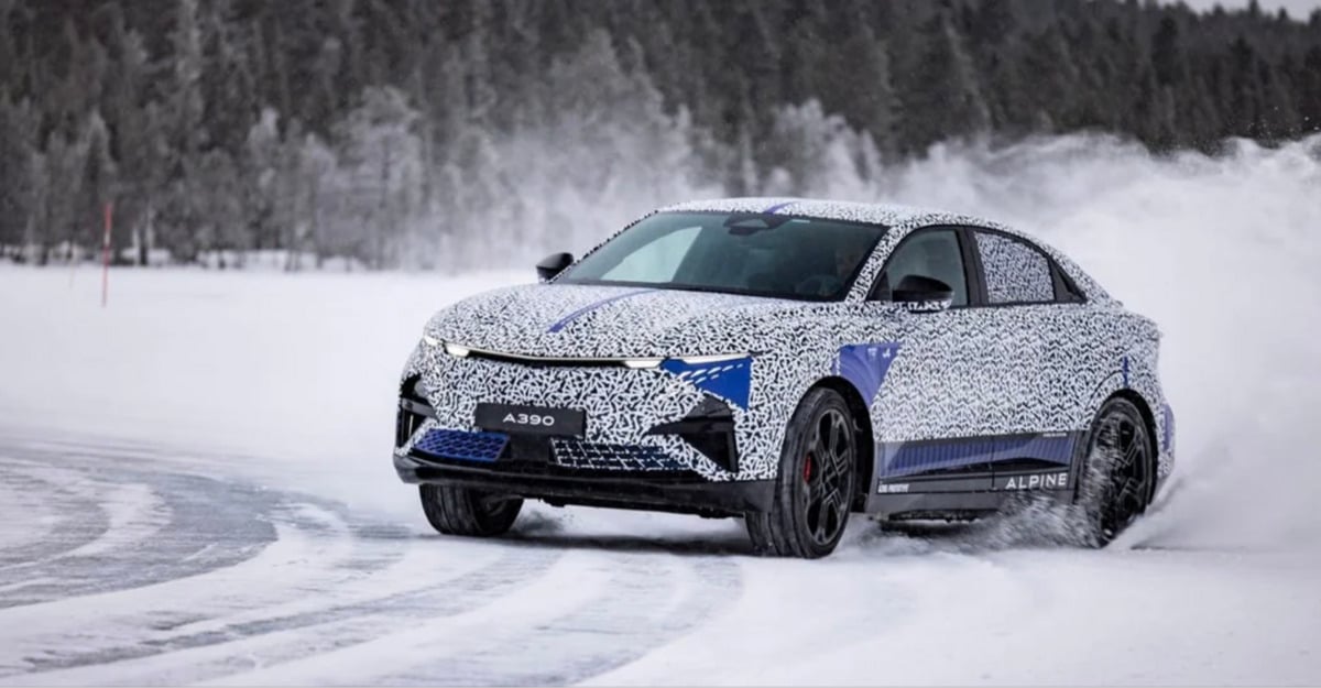A camouflaged Alpine A390 prototype car drives fast on a snowy track, kicking up snow, with a forest of bare trees in the background.