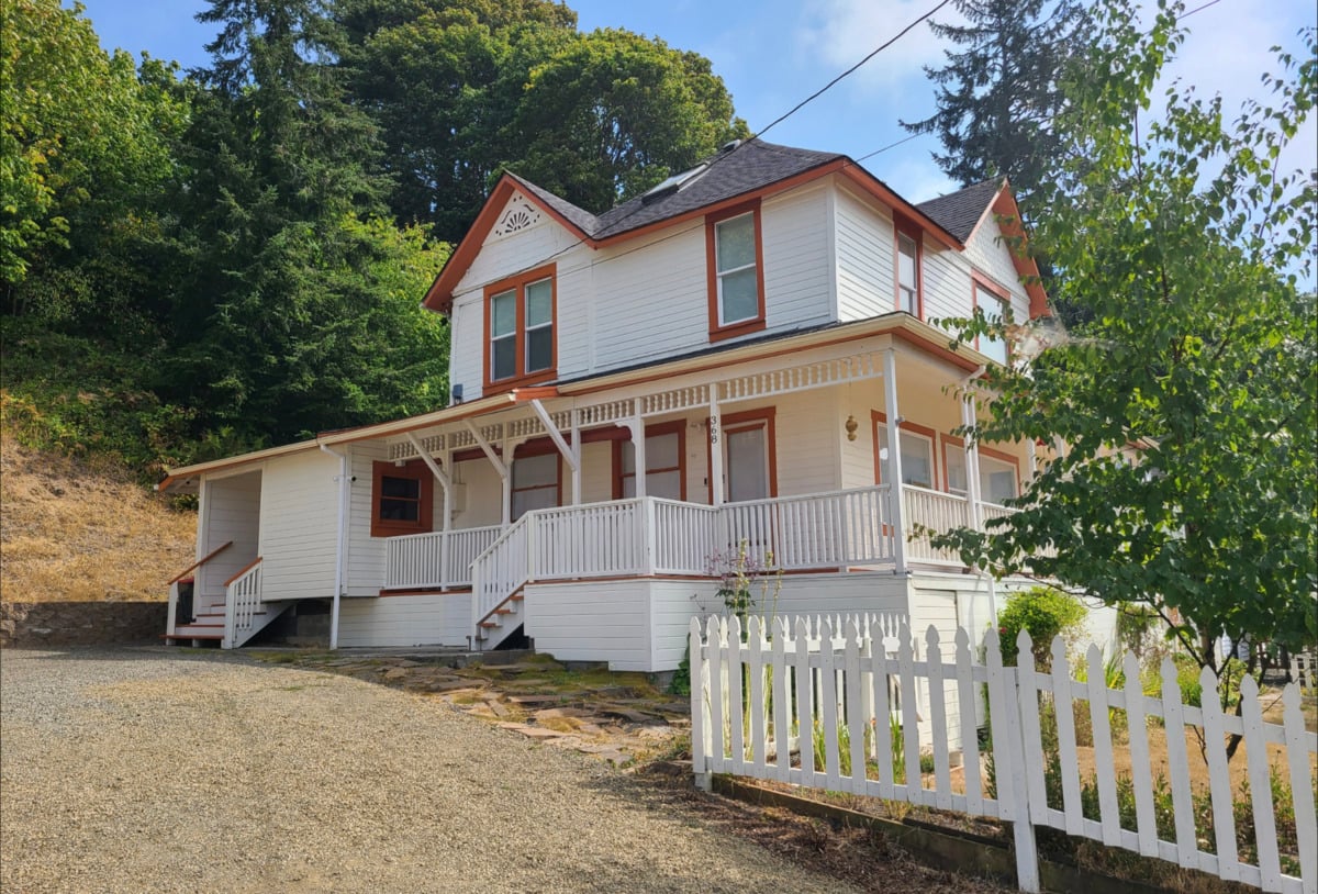 A two-story white house with a wraparound porch, red trim, and a white picket fence sits on a sloped gravel driveway, surrounded by trees and greenery.