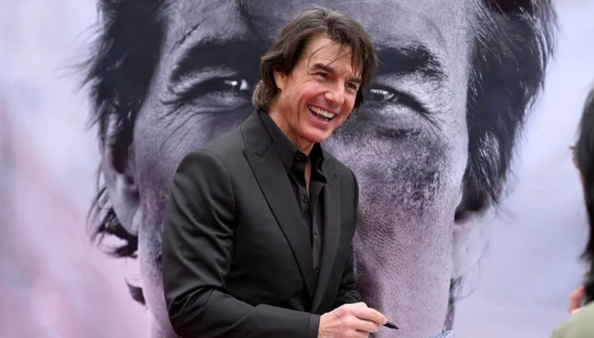 A man in a black suit smiles while signing an autograph in front of a large grayscale image of his own face displayed on a backdrop.