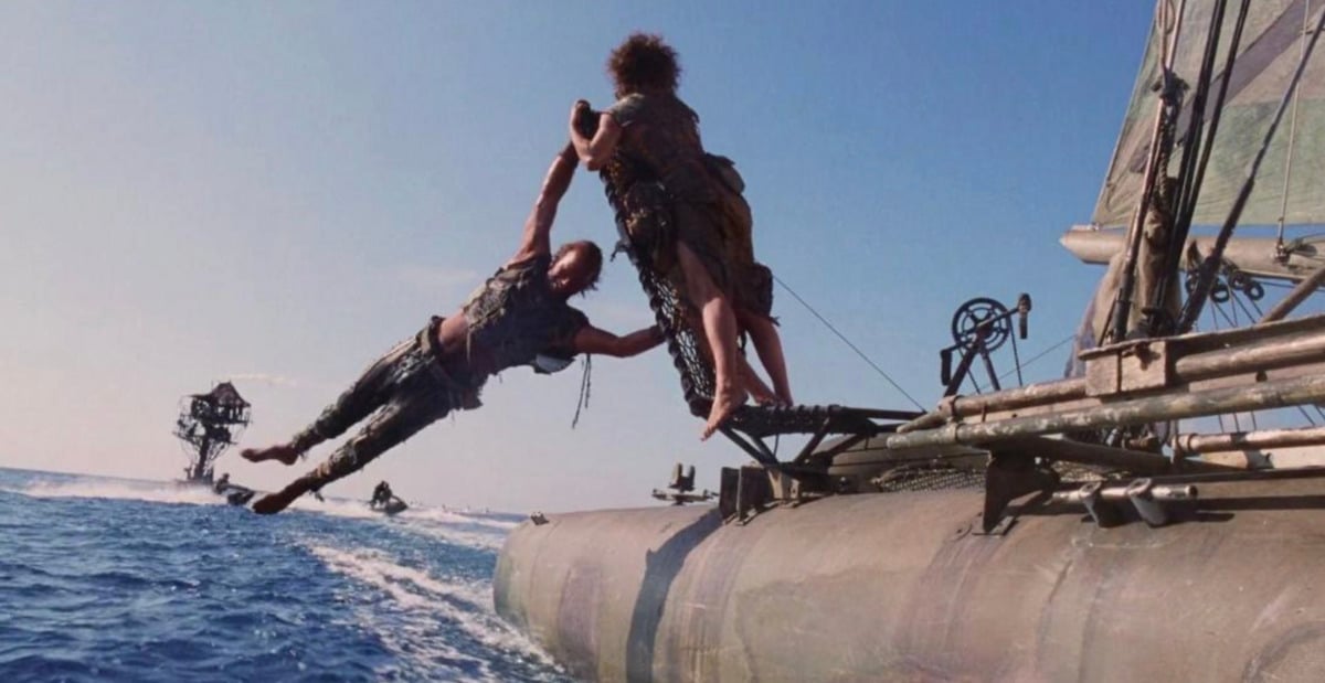 Two people in ragged clothes are on a sailboat at sea; one person is swinging another off the side of the boat while structures and boats are visible in the background under a clear blue sky.