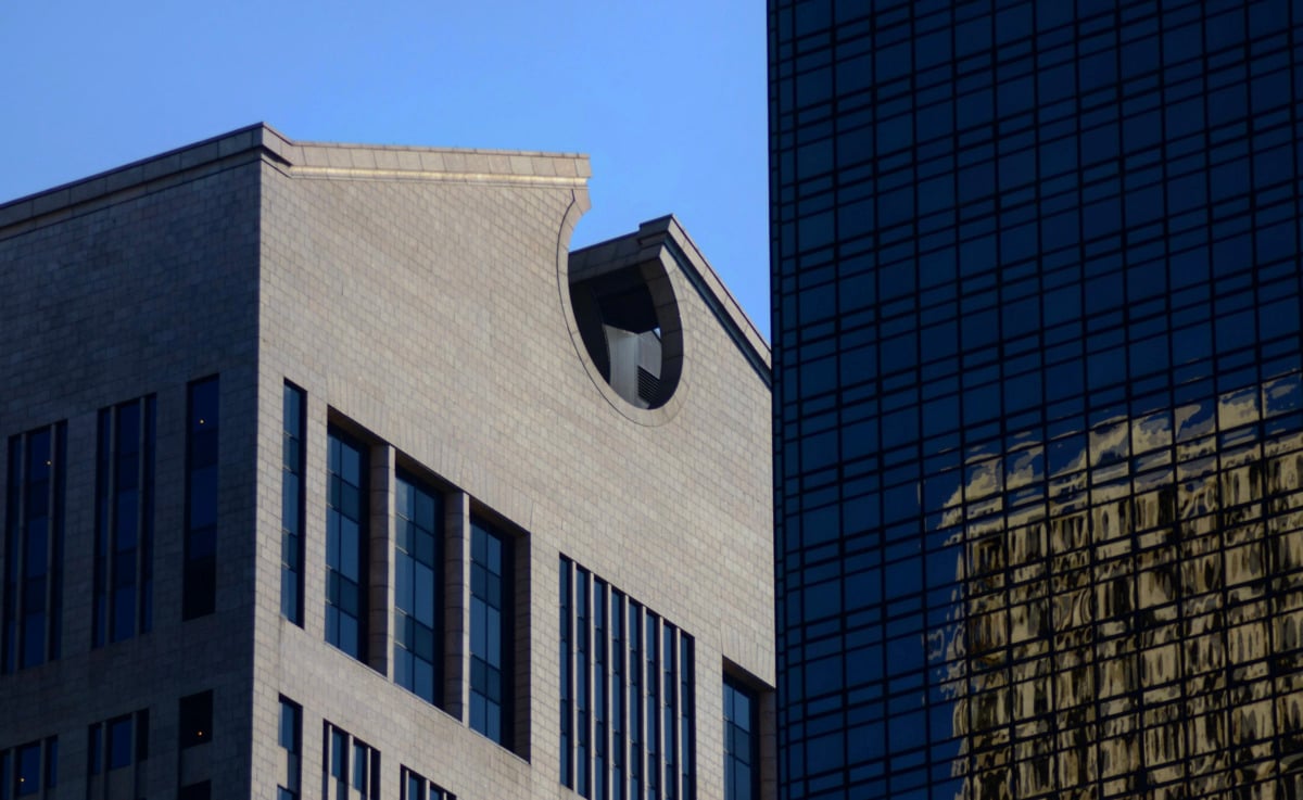 A light gray stone building with a distinctive semicircular cut-out at the roof stands beside a modern glass skyscraper reflecting another building against a clear blue sky.