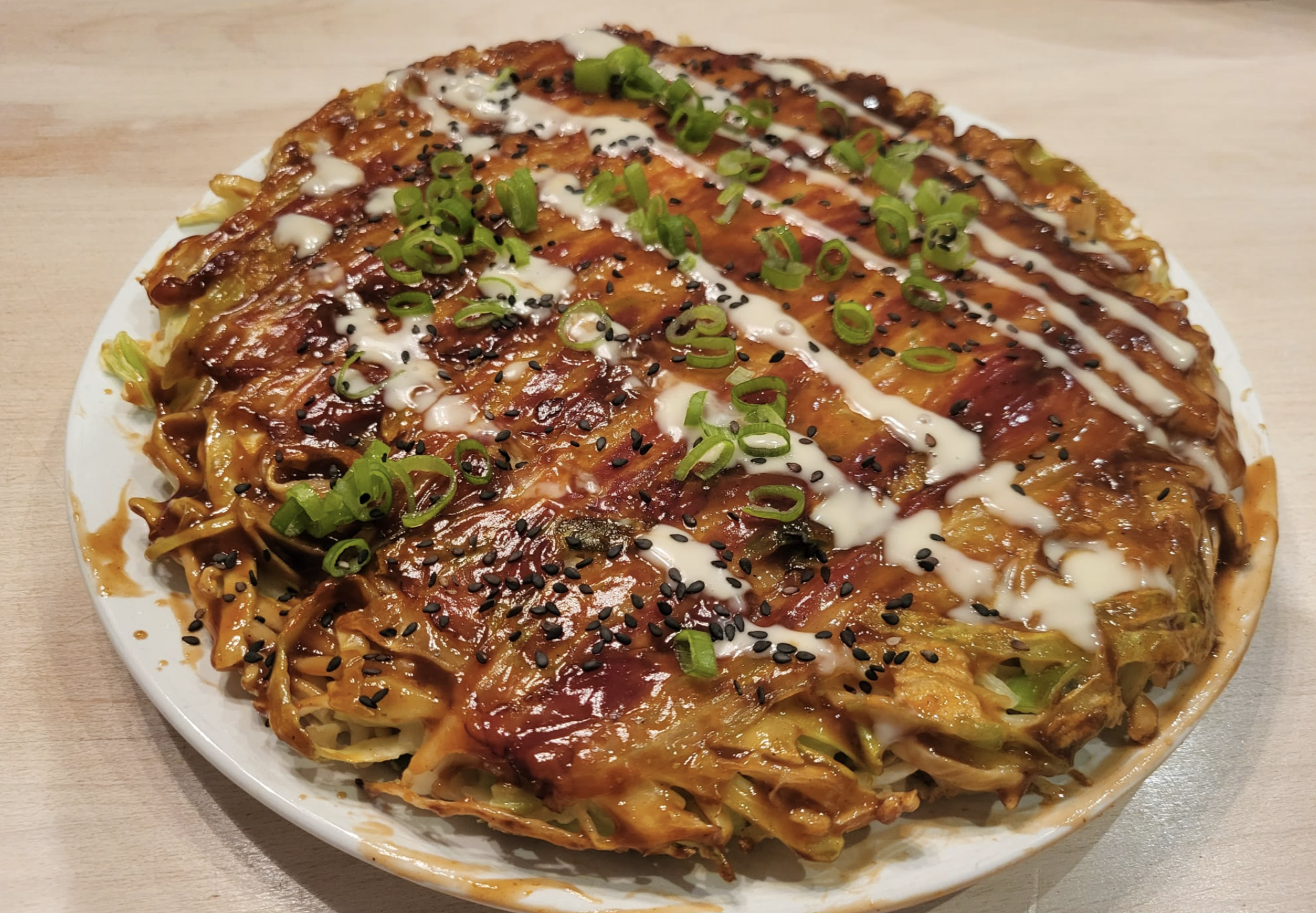 A large Japanese okonomiyaki pancake topped with savory sauces, drizzled mayonnaise, chopped green onions, and black sesame seeds, served on a white plate on a light-colored surface.