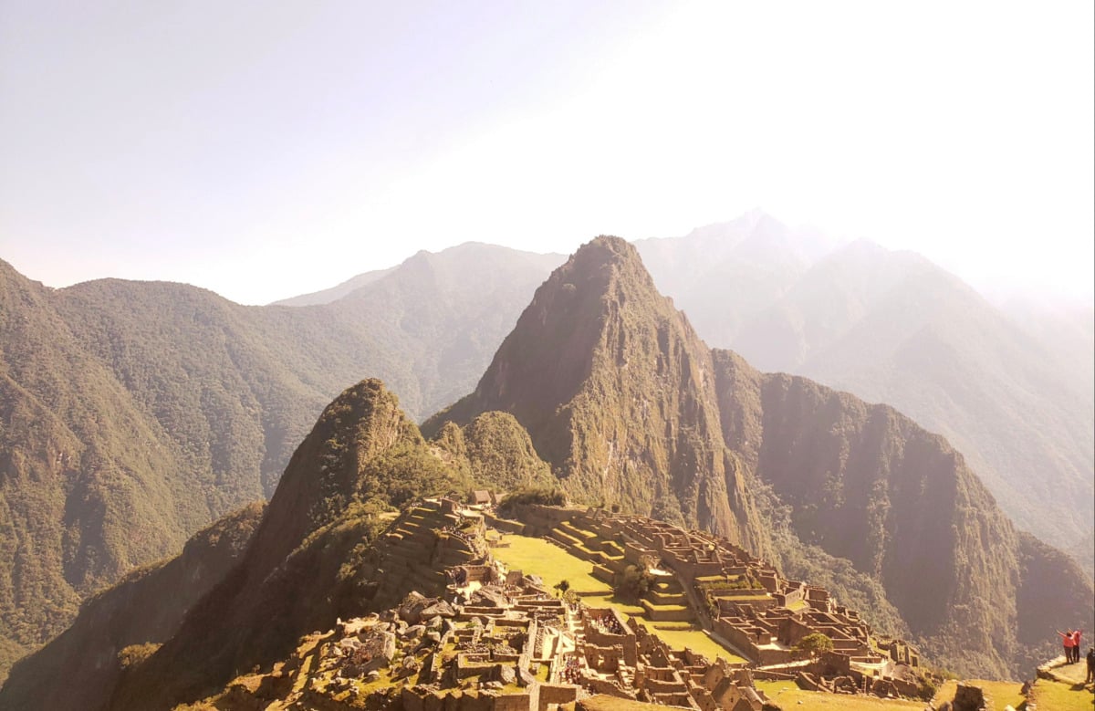 Ancient stone ruins of Machu Picchu sit atop a mountain ridge, surrounded by green, forested peaks under a bright, hazy sky.