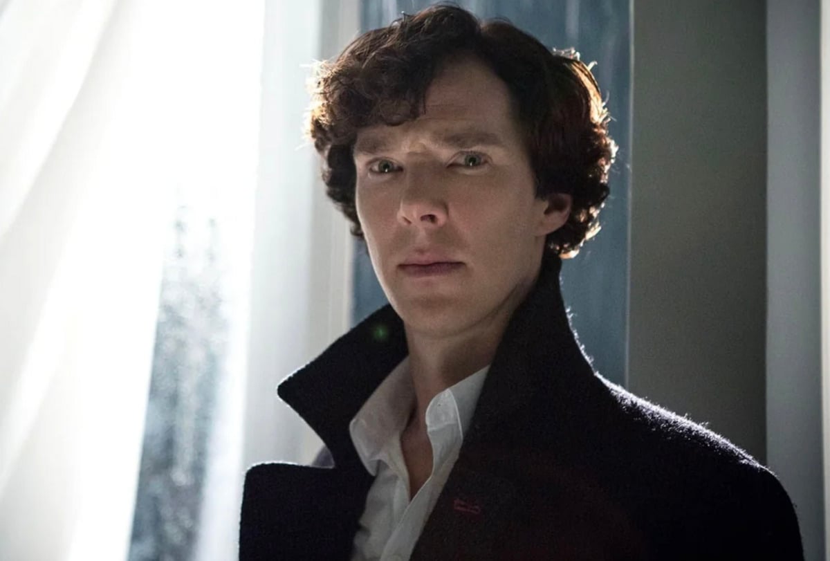 A man with curly brown hair, wearing a dark coat and white shirt, stands indoors near a window with sheer curtains, looking ahead with a serious expression. Light filters through the window, illuminating his face.