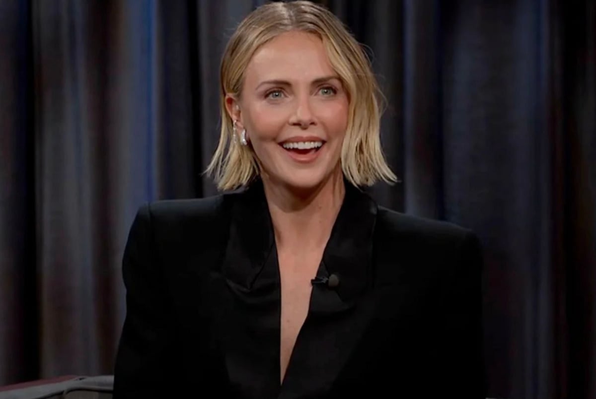 A woman with short blonde hair, wearing a black blazer, sits smiling during a talk show interview. Dark curtains are visible in the background.