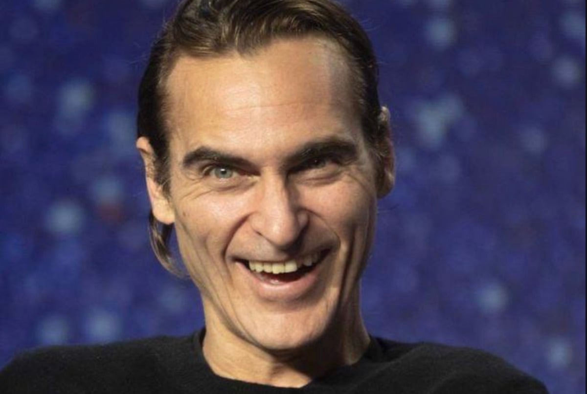 A man with slicked-back brown hair is smiling widely, wearing a black shirt. The background is blurred with blue and purple tones.