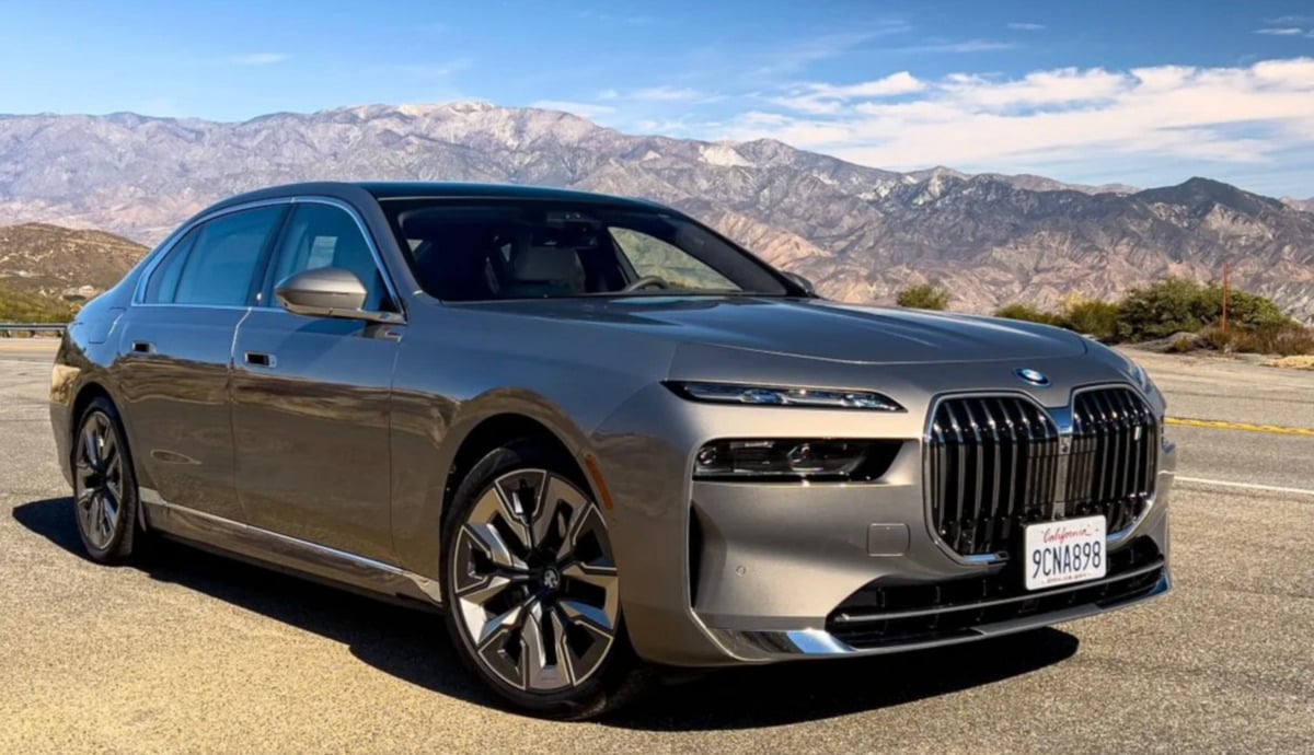 A silver BMW sedan is parked on the side of a road with mountains and a clear blue sky in the background. The car has a California license plate and modern, angular headlights.